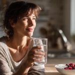 A woman looking calm and healthy in her sunlit kitchen, drinking a glass of water as a first step to find relief from medication-induced constipation.