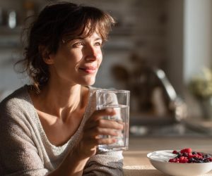 A woman looking calm and healthy in her sunlit kitchen, drinking a glass of water as a first step to find relief from medication-induced constipation.